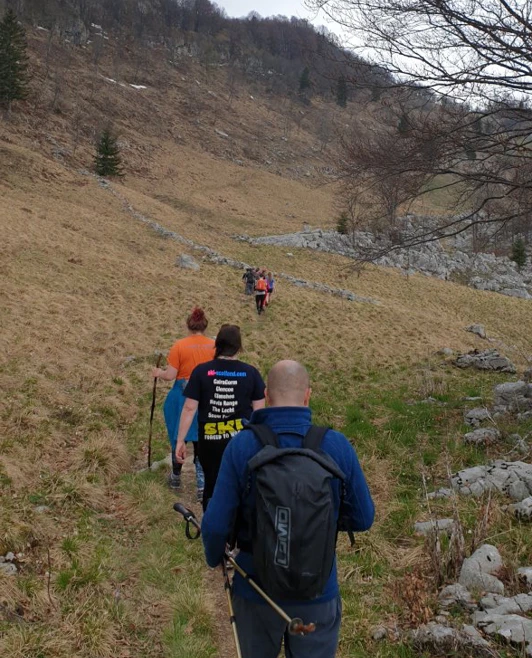 A group of hillwalkers in outdoor gear follow a narrow trail through open, hilly terrain with patches of woodland. A group of hillwalkers in outdoor gear follow a narrow trail through open, hilly terrain with patches of woodland.